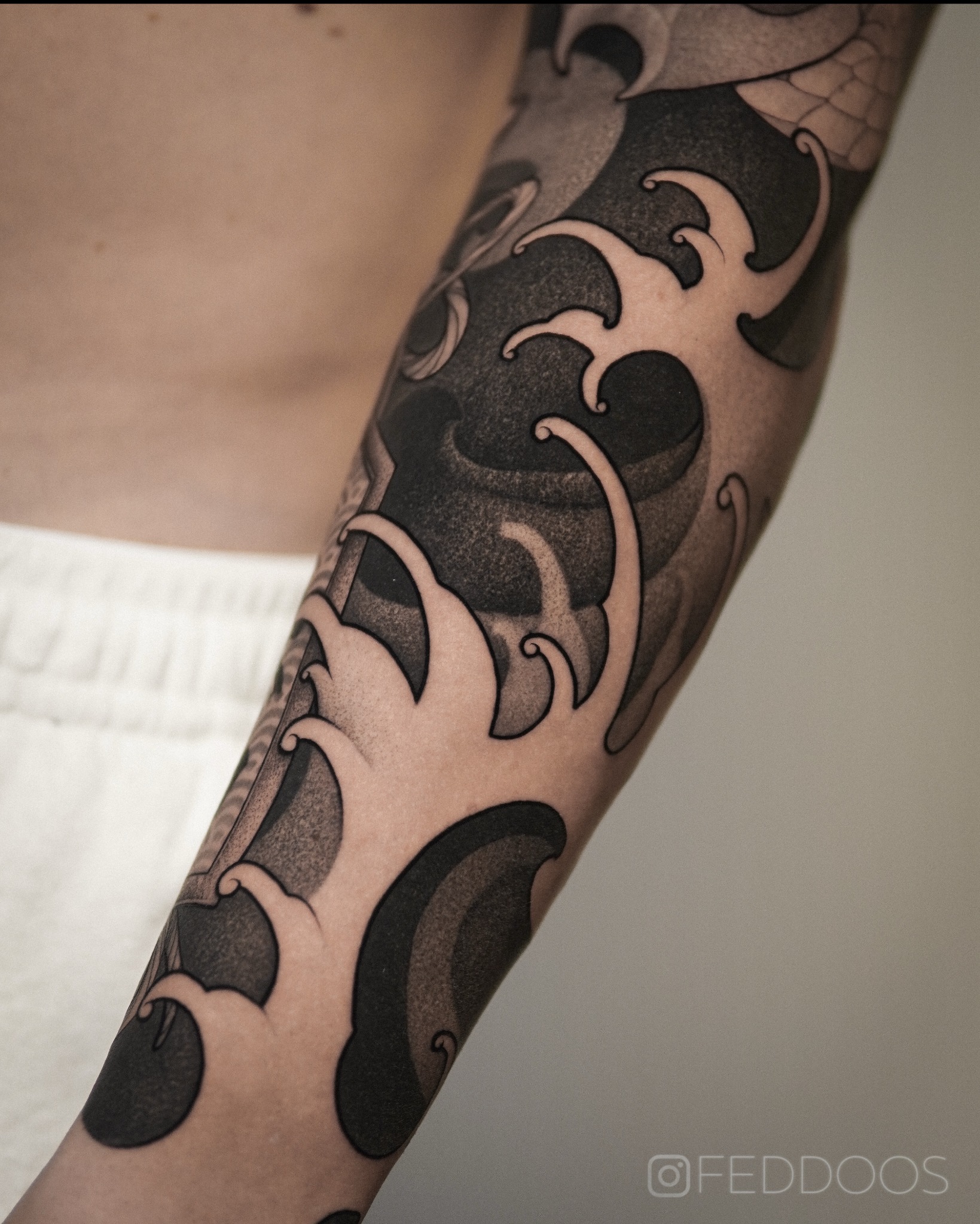 Ramię z czarno-białym tatuażem w stylu japońskim, przedstawiającym fale i smoka. Delikatne cieniowanie, widoczny podpis artysty w prawym dolnym rogu.