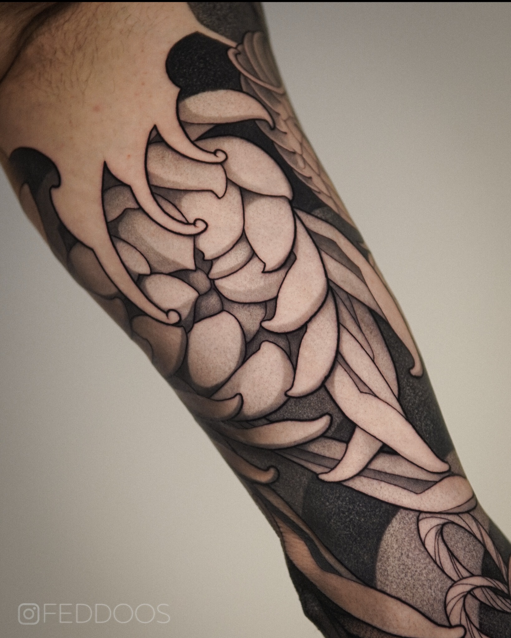 Czarno-biały tatuaż na ramieniu z motywem kwiatowym i elementami abstrakcyjnymi, wykonany z dbałością o detale i cieniowanie, widoczne logo wykonawcy w rogu.