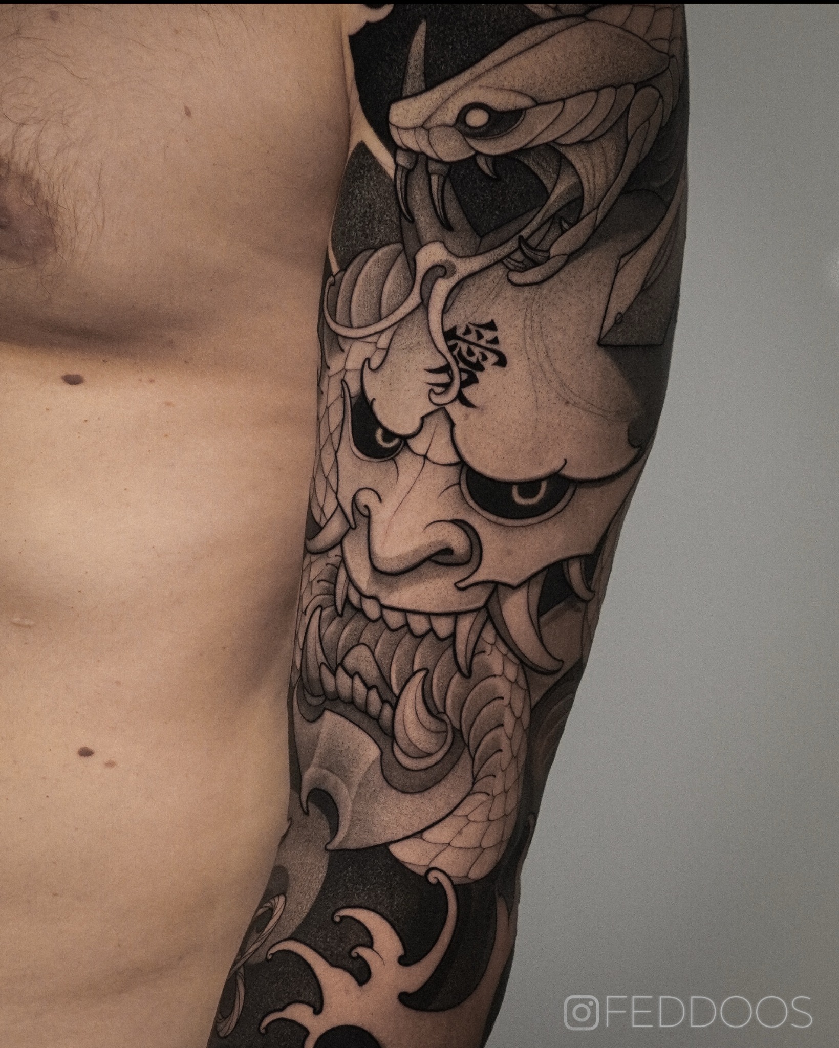 Czarno-biały tatuaż na ramieniu mężczyzny przedstawiający demona Hannya z wężem i motywem fal, wykonany z dbałością o detale i cieniowanie.