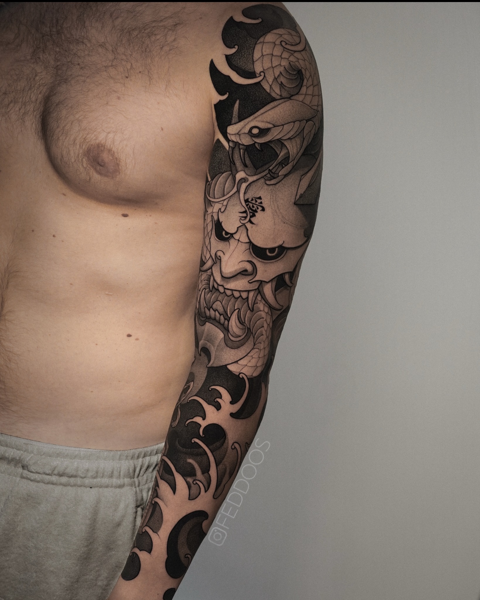 Męskie ramię z czarno-szarym tatuażem w stylu japońskim: maska Hannya, wąż i fale. Widoczny fragment klatki piersiowej i szare spodnie dresowe. Sygnatura tatuażysty.