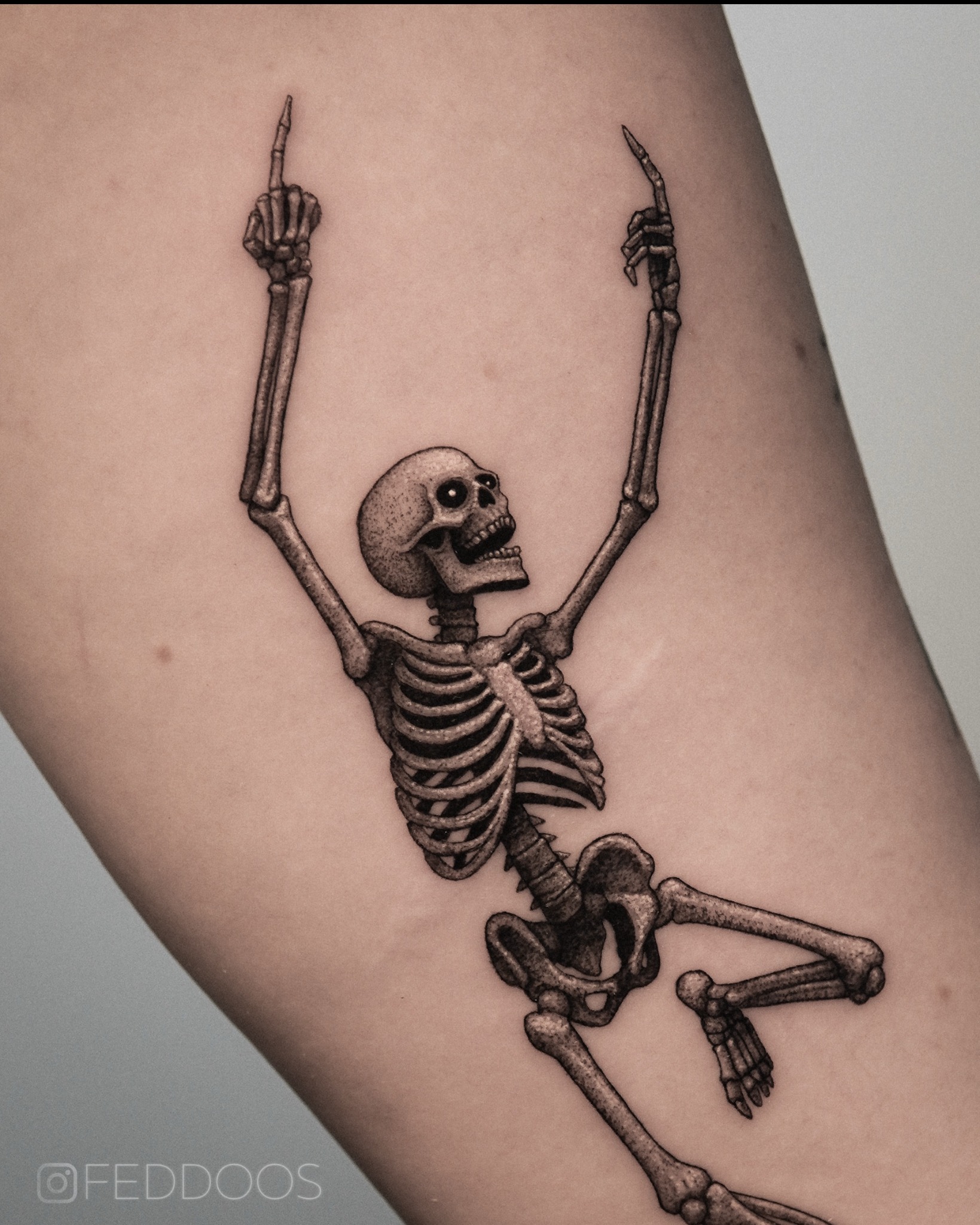 Czarno-szary tatuaż na ramieniu przedstawiający szkielet z uniesionymi rękami i wyciągniętymi palcami wskazującymi. Widoczne detale cieniowania i anatomiczne szczegóły.