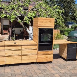 WOOD METAL ART SPÓŁKA Z OGRANICZONĄ ODPOWIEDZIALNOŚCIĄ - Mobilna kuchnia outdoorowa z drewna na kółkach, z piekarnikiem i grillem, na tle brukowanej nawierzchni i zieleni. Elementy łatwe w demontażu.