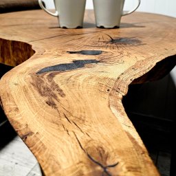 WOOD METAL ART SPÓŁKA Z OGRANICZONĄ ODPOWIEDZIALNOŚCIĄ - Blat stołu z litego drewna dębowego z żywicą epoksydową, wykończony na wysoki połysk. Na blacie stoją dwie białe filiżanki. Widoczne słoje i naturalne krawędzie drewna.