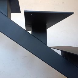 WOOD METAL ART SPÓŁKA Z OGRANICZONĄ ODPOWIEDZIALNOŚCIĄ - Minimalistyczne, czarne schody metalowe z płaskimi stopniami, widziane z dołu pod kątem, na tle białej ściany z delikatną fakturą. Widoczne mocowania stopni.