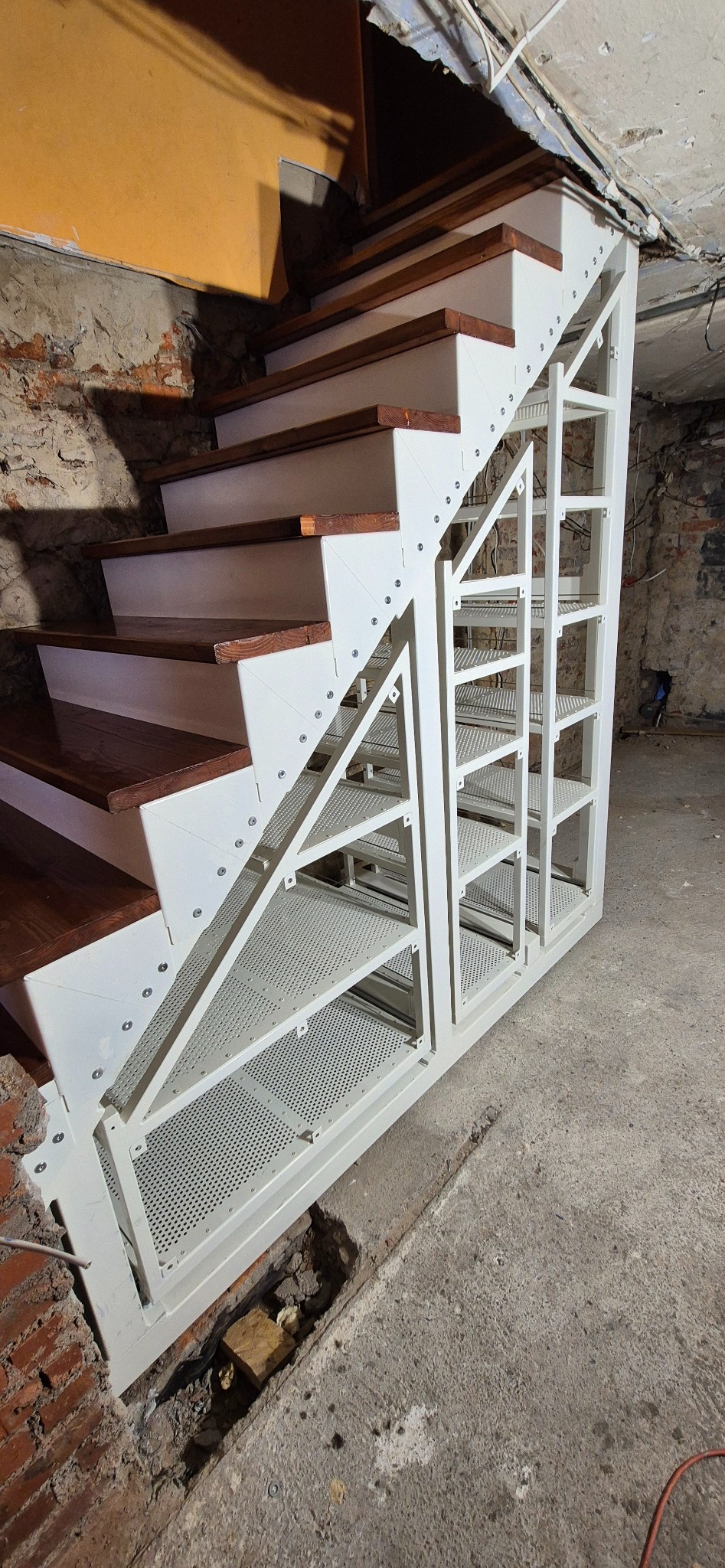 Schody z metalową konstrukcją i drewnianymi stopniami, zintegrowane z systemem wysuwanych szafek. Biała rama, brązowe stopnie, surowe wykończenie ścian.