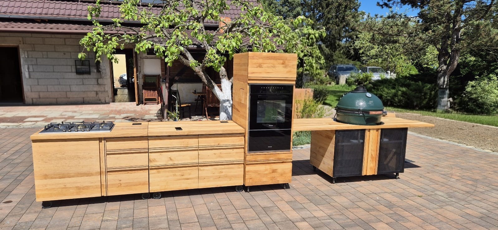 Mobilna kuchnia outdoorowa z drewna na kółkach, z piekarnikiem i grillem, na tle brukowanej nawierzchni i zieleni. Elementy łatwe w demontażu.