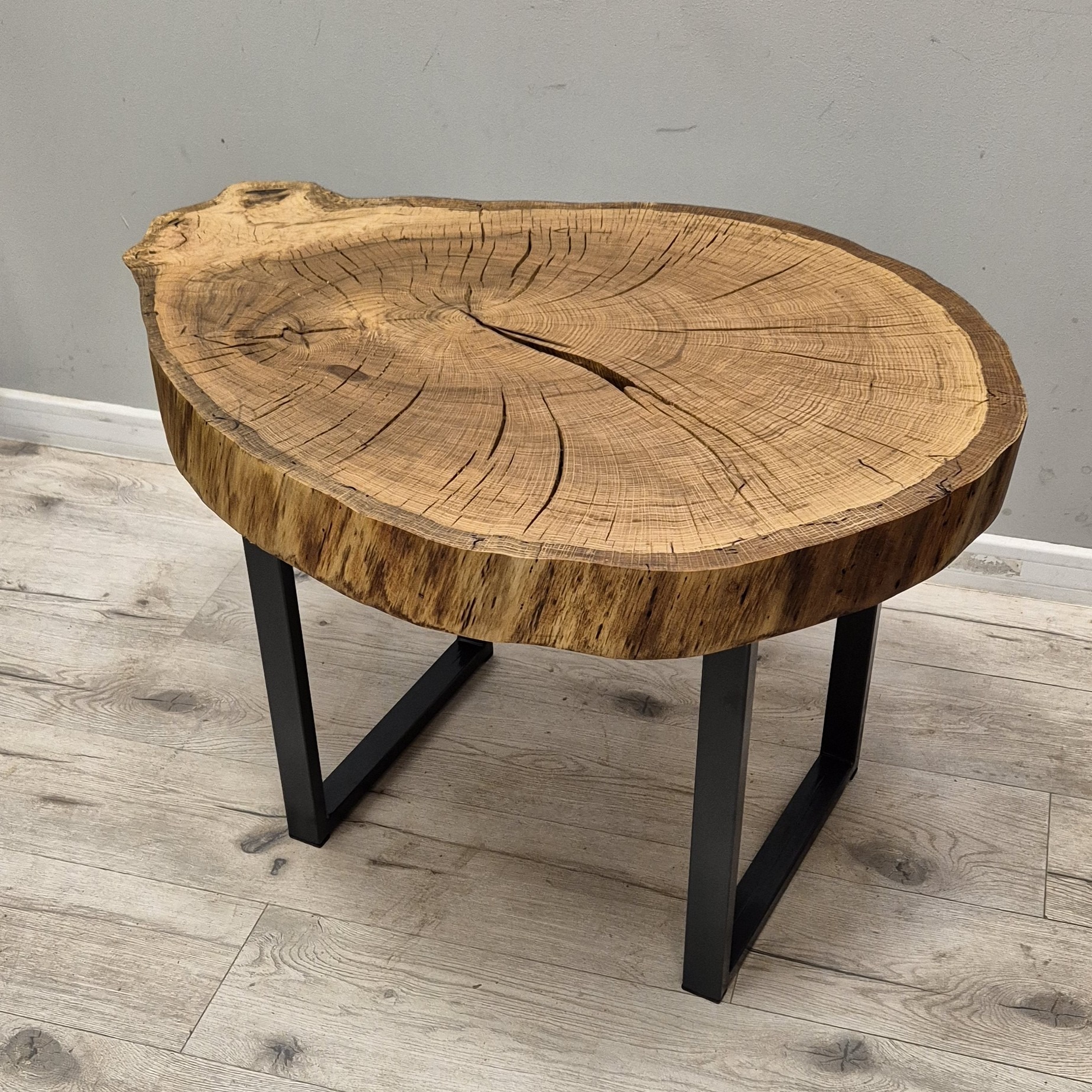 Stolik kawowy z blatem z plastra drewna o wyraźnych słojach, osadzony na czarnych, metalowych nogach w kształcie litery U. Minimalistyczny design na tle szarej ściany i drewnianej podłogi.