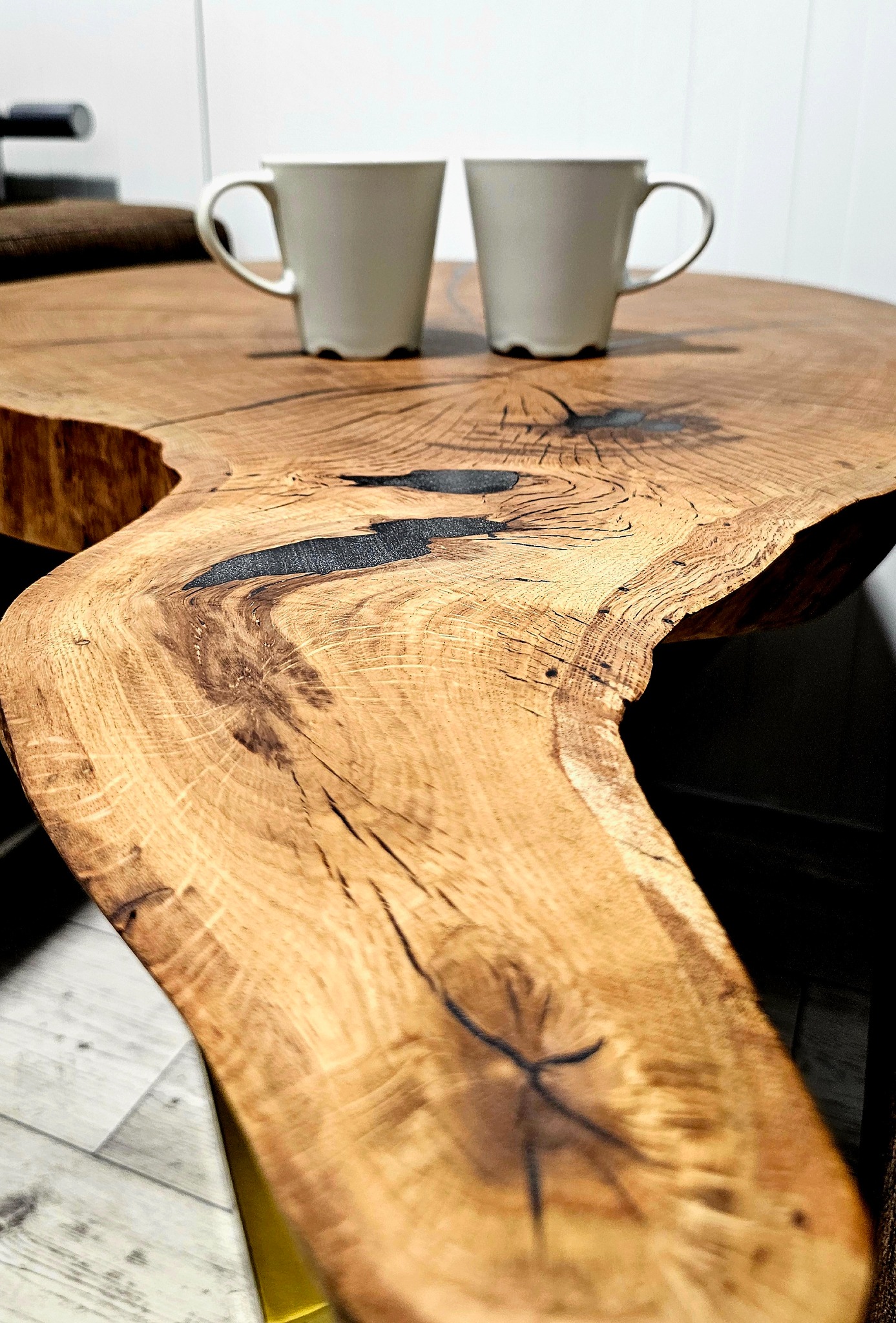 Blat stołu z litego drewna dębowego z żywicą epoksydową, wykończony na wysoki połysk. Na blacie stoją dwie białe filiżanki. Widoczne słoje i naturalne krawędzie drewna.