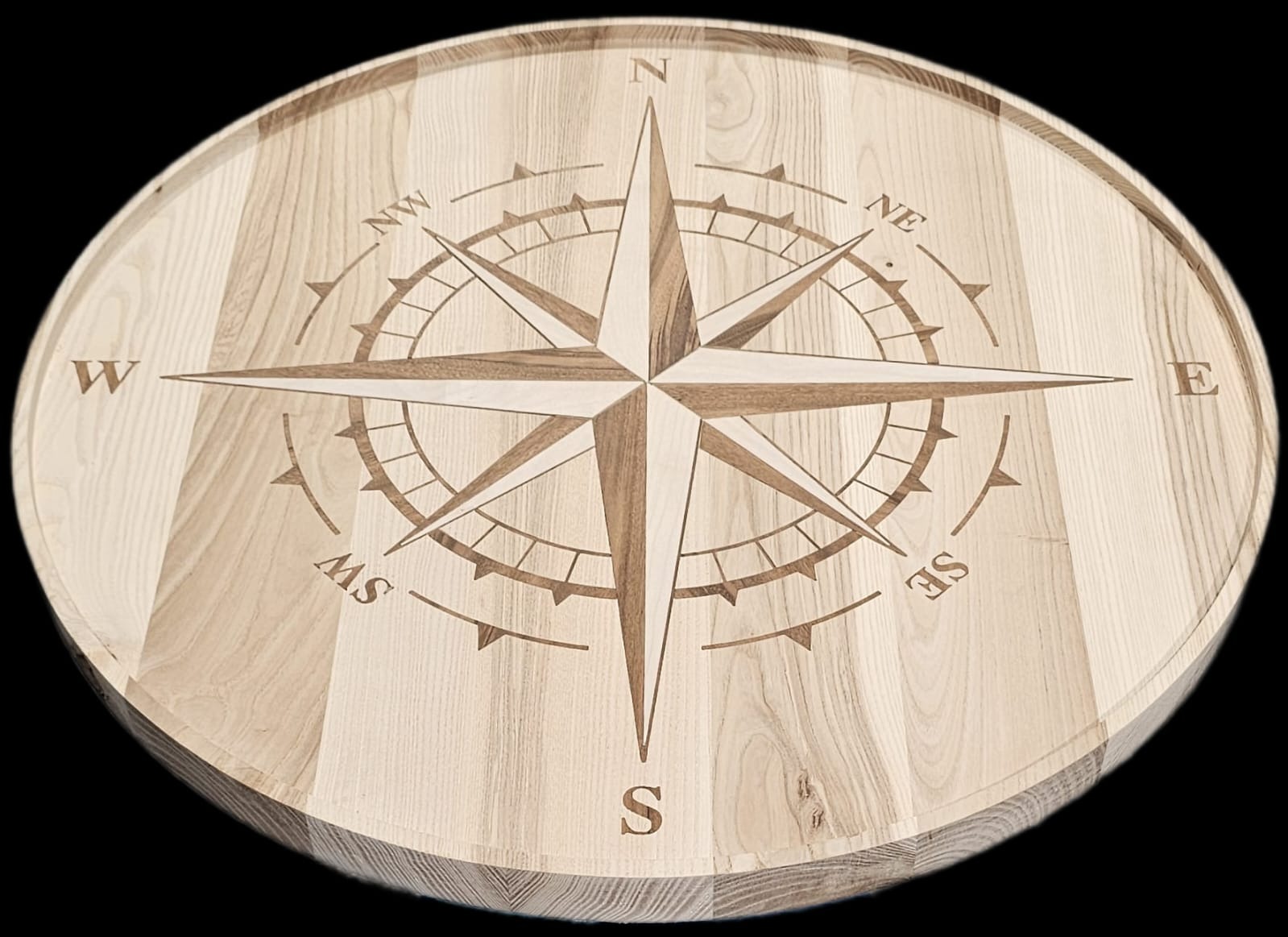 Okrągły, drewniany blat z wygrawerowaną różą kompasową. Jasne drewno z wyraźnym usłojeniem. Precyzyjne detale grawerunku. Widoczne oznaczenia kierunków świata.