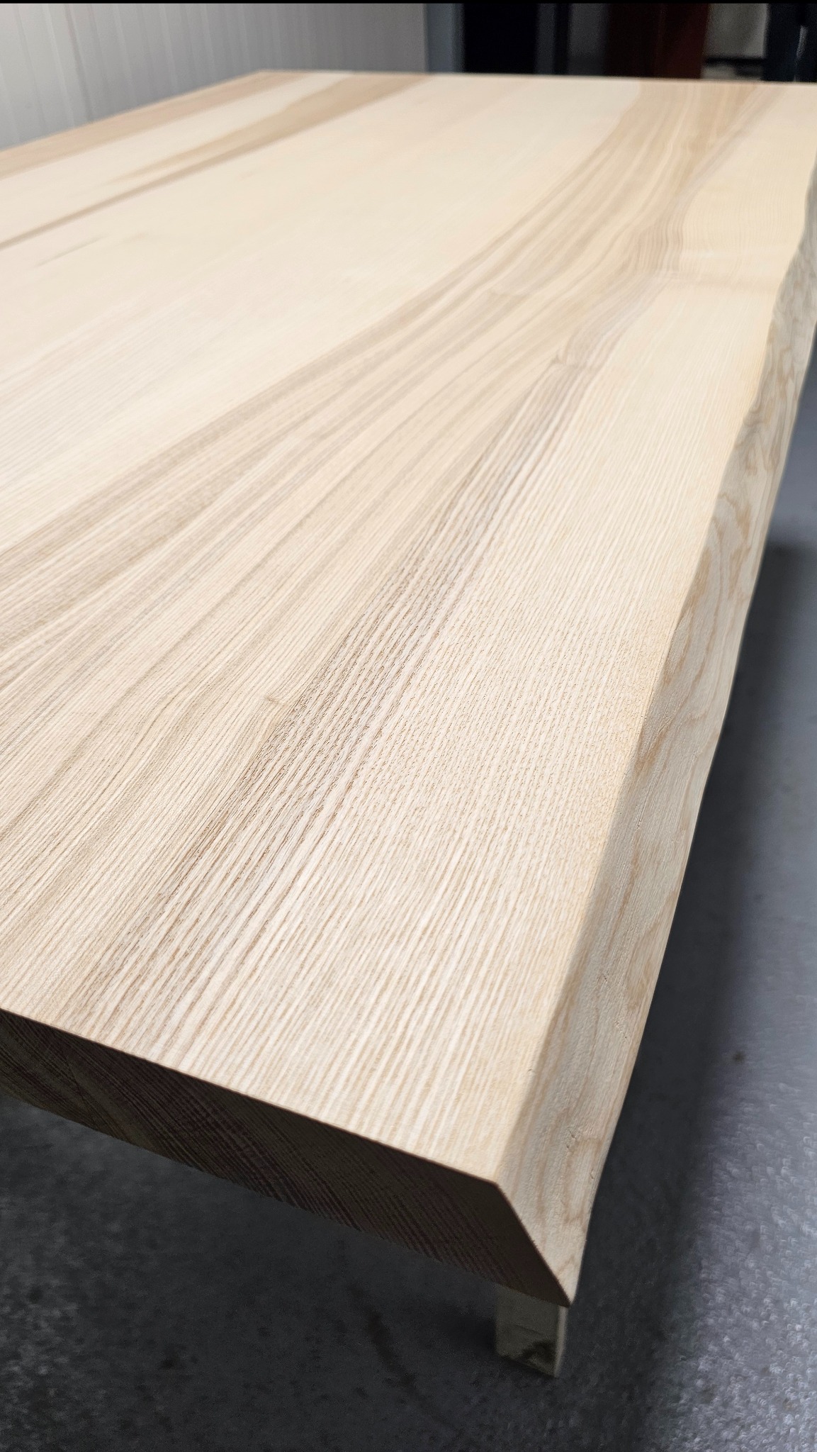 Blat stołu z jasnego drewna o nieregularnej krawędzi, ukazujący naturalny rysunek słojów. Minimalistyczne nóżki. Wykończenie matowe.