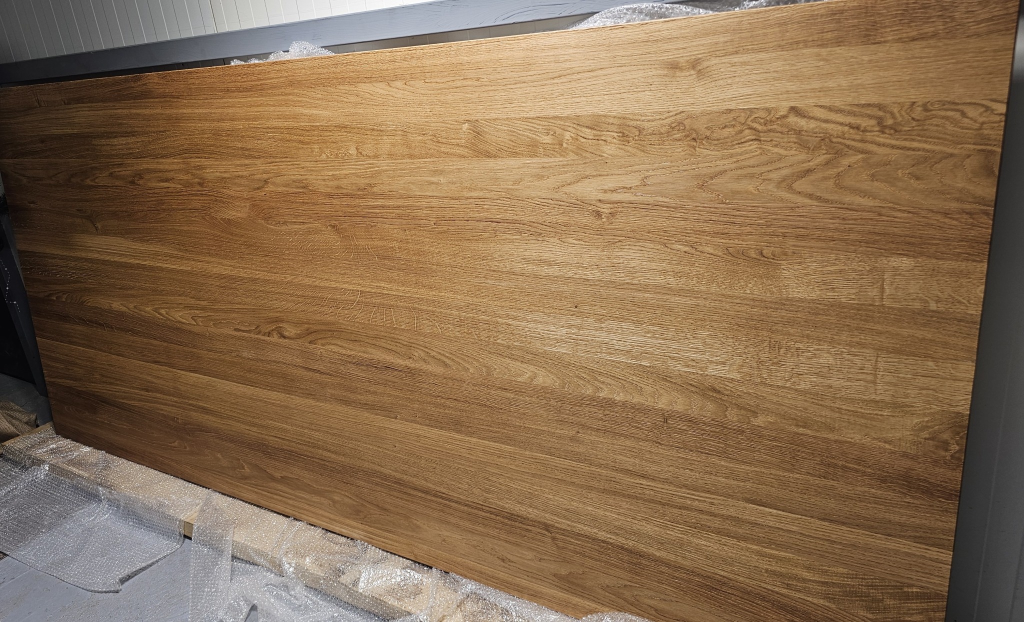 Blat z litego drewna dębowego o wyraźnym usłojeniu, zabezpieczony folią bąbelkową, gotowy do montażu. Ciepłe oświetlenie podkreśla naturalny rysunek drewna.