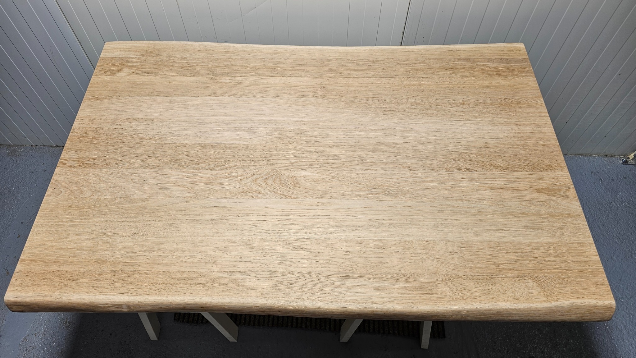 Blat stołu z jasnego drewna o nieregularnych krawędziach, widoczny z góry, wsparty na białych nogach. Tło stanowi szara ściana z panelami.