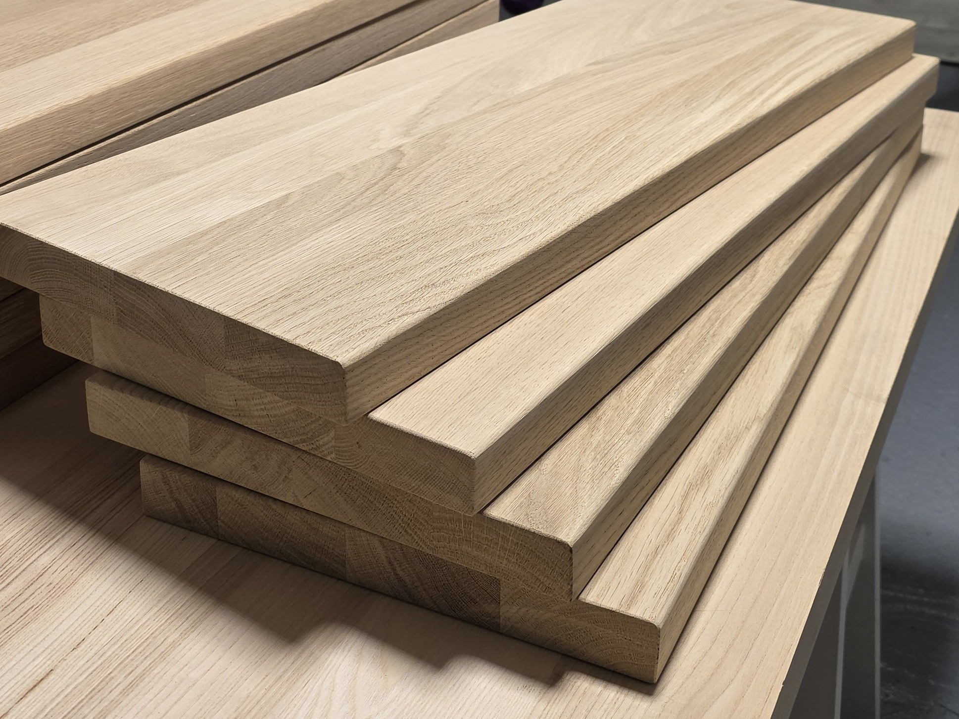 Stos jasnych, drewnianych stopni schodowych z widocznym usłojeniem, ułożonych jeden na drugim. Elementy przygotowane do montażu, o gładkiej powierzchni i zaokrąglonych krawędziach.