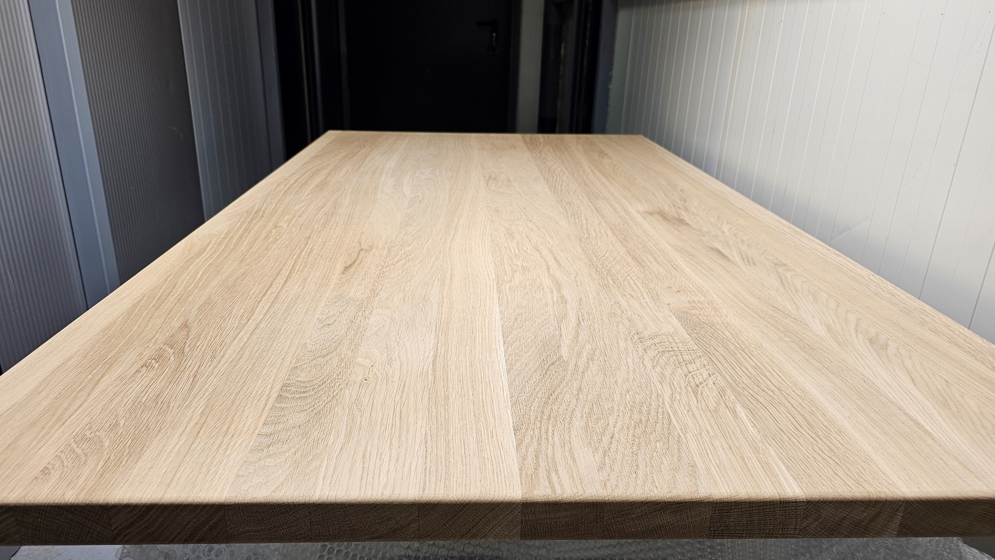 Jasny, dębowy blat z wyraźnym usłojeniem, sfotografowany pod kątem, ukazujący jego strukturę i wykończenie. Tło stanowią pionowe panele w odcieniach szarości i bieli.