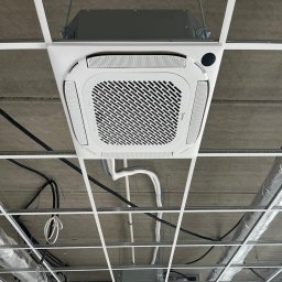 CoolPro Bogumił Brzozowski - Kasetonowa jednostka klimatyzacji Midea zamontowana w suficie podwieszanym, widok z dołu, z widocznymi przewodami i elementami konstrukcyjnymi sufitu.
