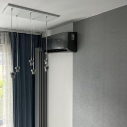 CoolPro Bogumił Brzozowski - Nowoczesny, czarny klimatyzator ścienny z wyświetlaczem temperatury, zamontowany w szarym pokoju z grzejnikiem i lampą wiszącą. Widok z dołu.