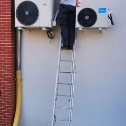 CoolPro Bogumił Brzozowski - Instalator na drabinie montuje dwie jednostki klimatyzacji Midea na elewacji budynku z ceglanym narożnikiem. Widoczne przewody i elementy montażowe.