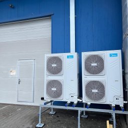 CoolPro Bogumił Brzozowski - Dwa białe klimatyzatory Midea zamontowane na metalowej konstrukcji przed niebiesko-białym budynkiem. Widoczne rury i kostka brukowa. Chłodne oświetlenie.