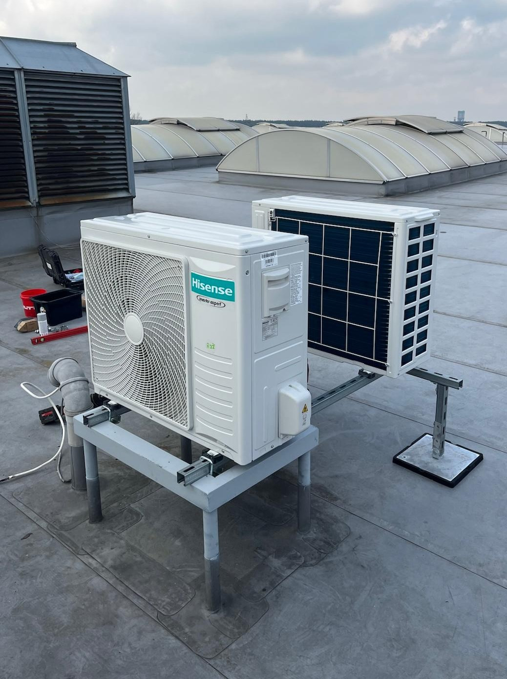 Dwa białe klimatyzatory Hisense na dachu budynku, zamontowane na szarych metalowych konstrukcjach wsporczych. W tle świetliki dachowe i narzędzia.
