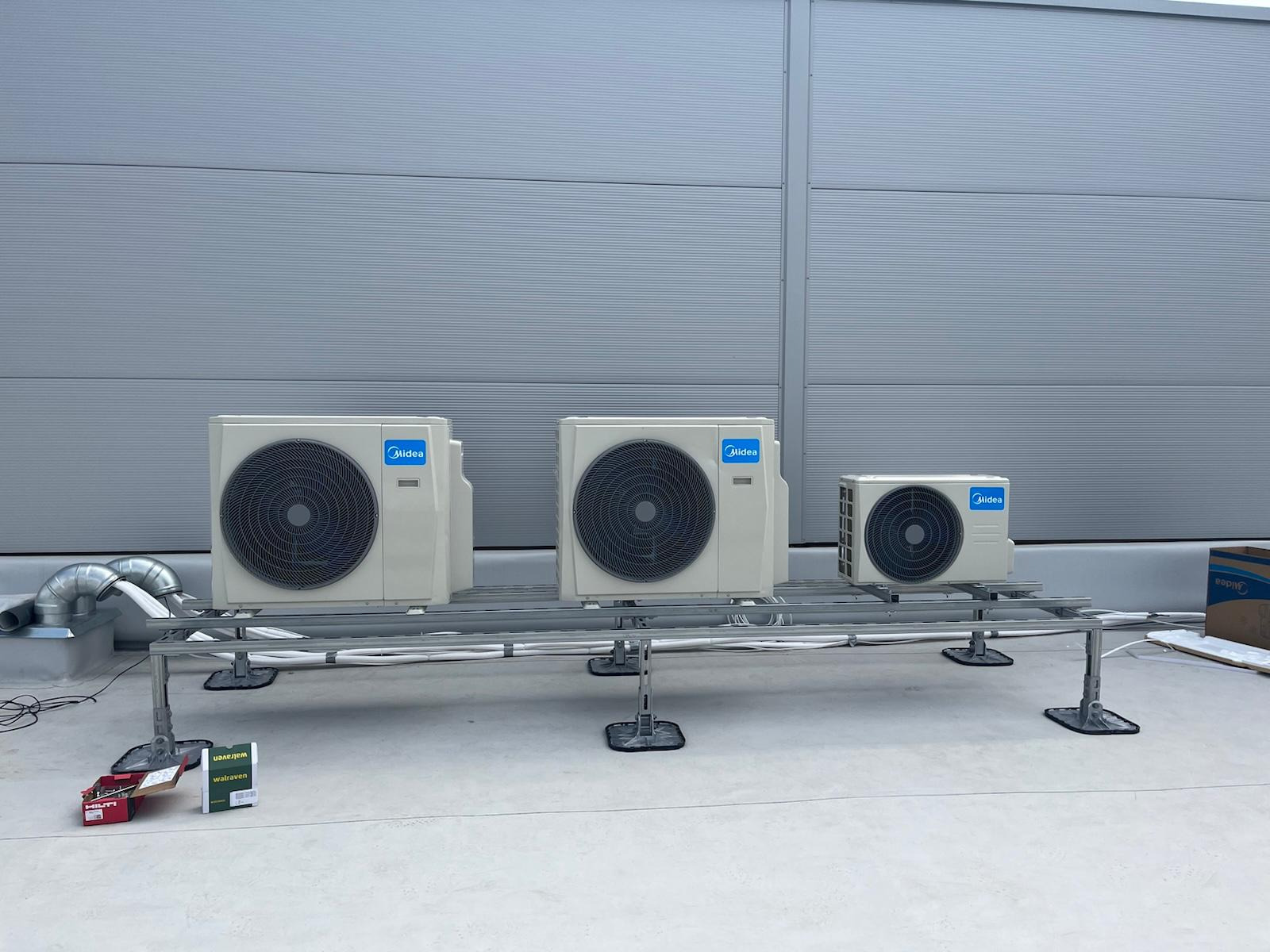 Trzy klimatyzatory Midea na metalowej konstrukcji na dachu budynku o szarej, panelowej elewacji. Widoczne przewody i akcesoria montażowe.