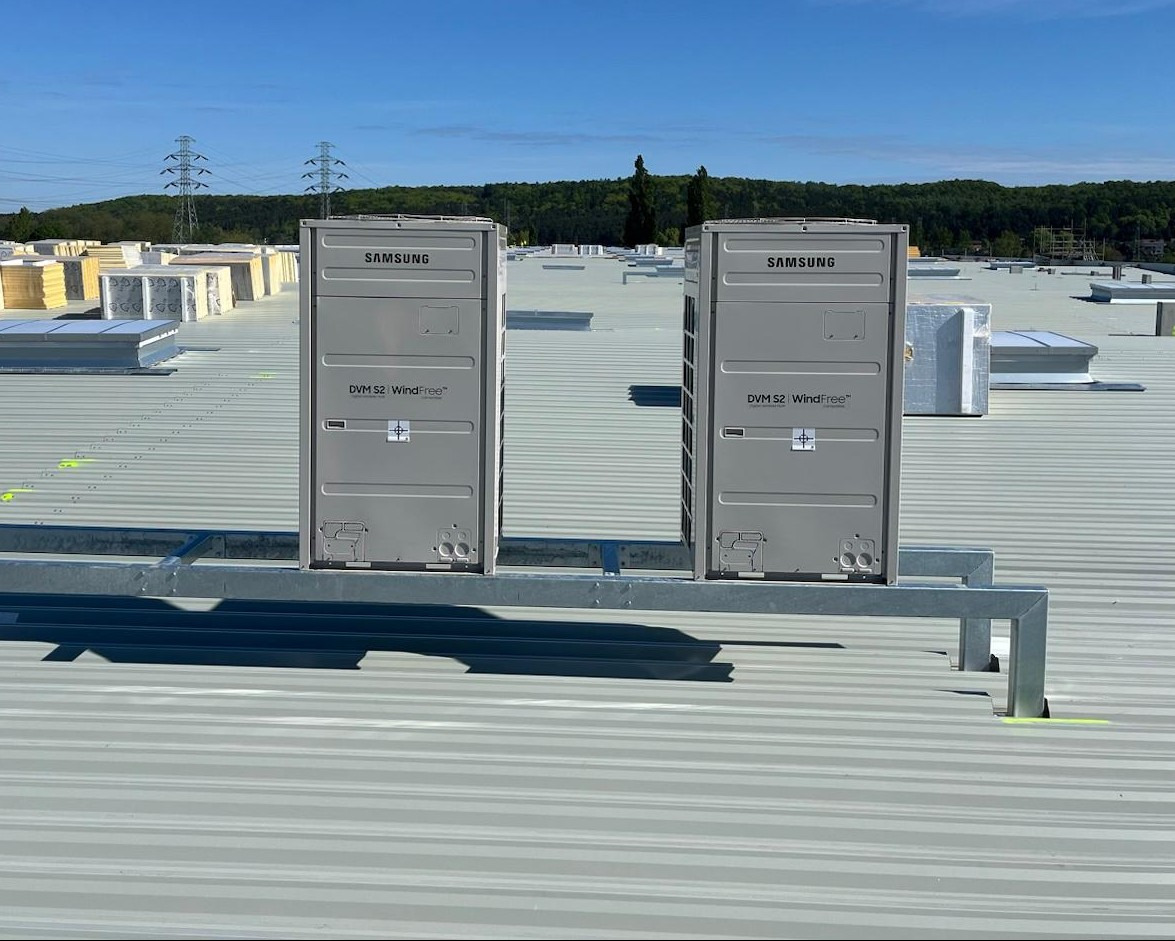 Dwa szare, identyczne klimatyzatory Samsung DVM S2 WindFree™ na dachu budynku komercyjnego, zamontowane na metalowej konstrukcji, z widocznymi elementami instalacji.