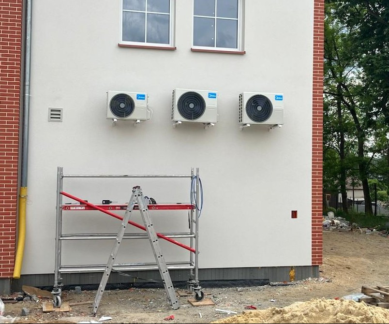 Trzy klimatyzatory Midea zamontowane na białej ścianie budynku z ceglanym narożnikiem. Na pierwszym planie rusztowanie i drabina, sugerujące trwający montaż.