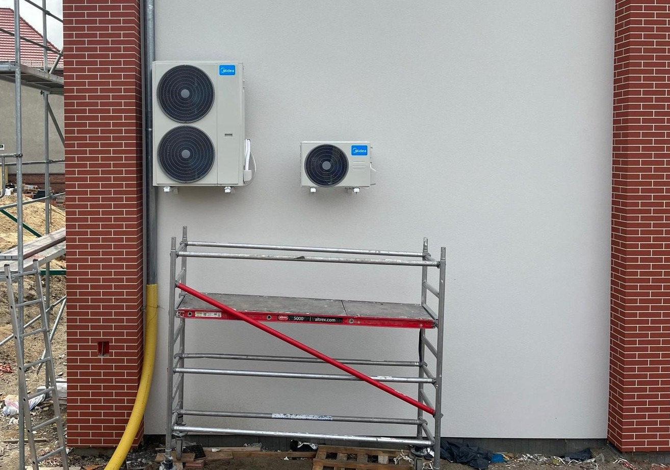 Dwa klimatyzatory Midea zamontowane na białej ścianie budynku z ceglanymi elementami dekoracyjnymi. Widoczne rusztowanie.