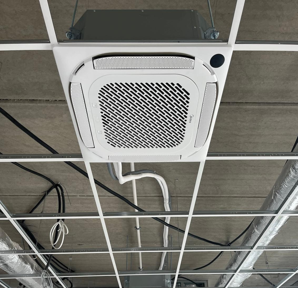 Kasetonowa jednostka klimatyzacji Midea zamontowana w suficie podwieszanym, widok z dołu, z widocznymi przewodami i elementami konstrukcyjnymi sufitu.