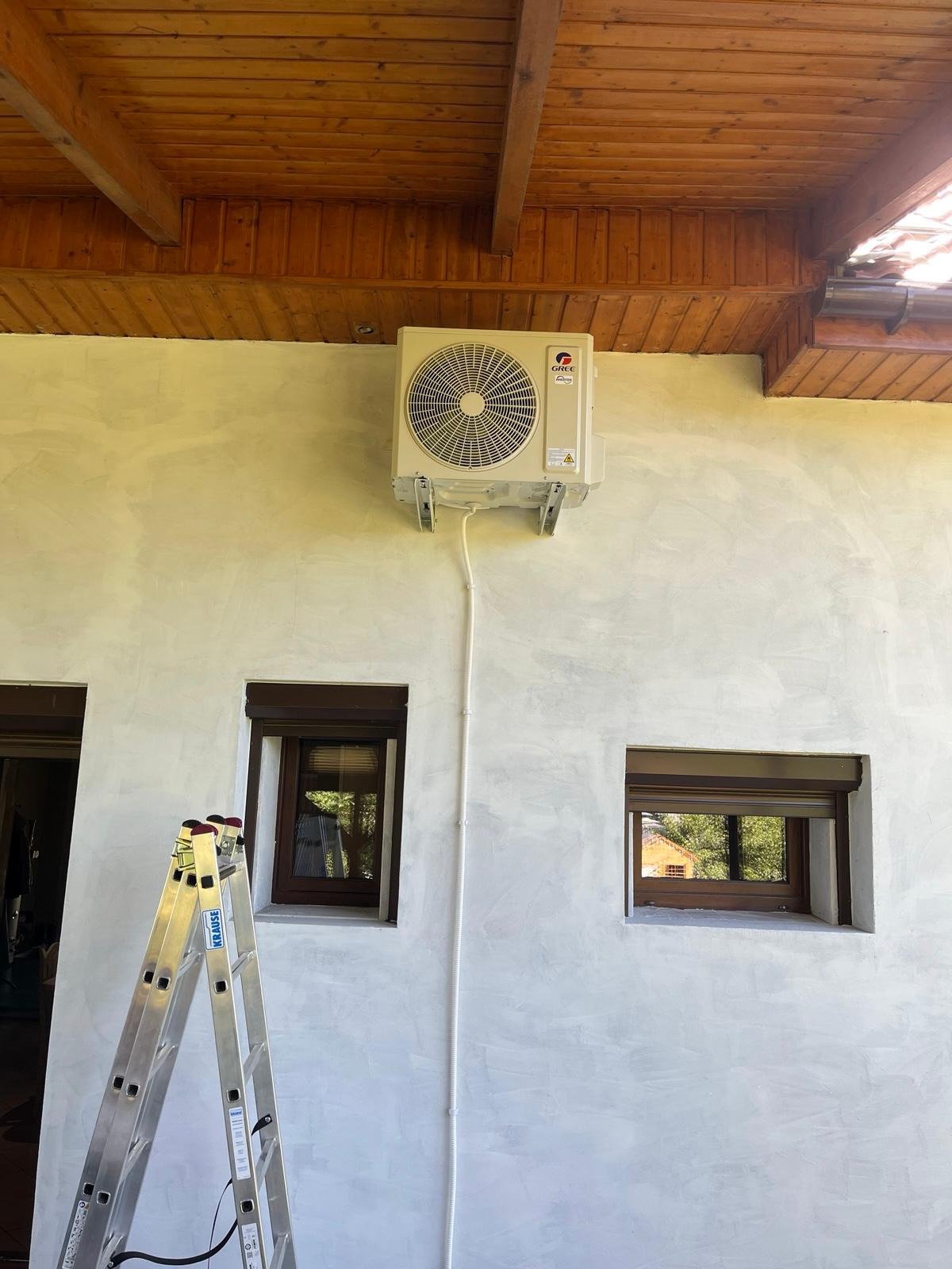 Zamontowana jednostka zewnętrzna klimatyzacji marki Gree na elewacji budynku z widoczną drabinką i dwoma oknami z brązowymi ramami.