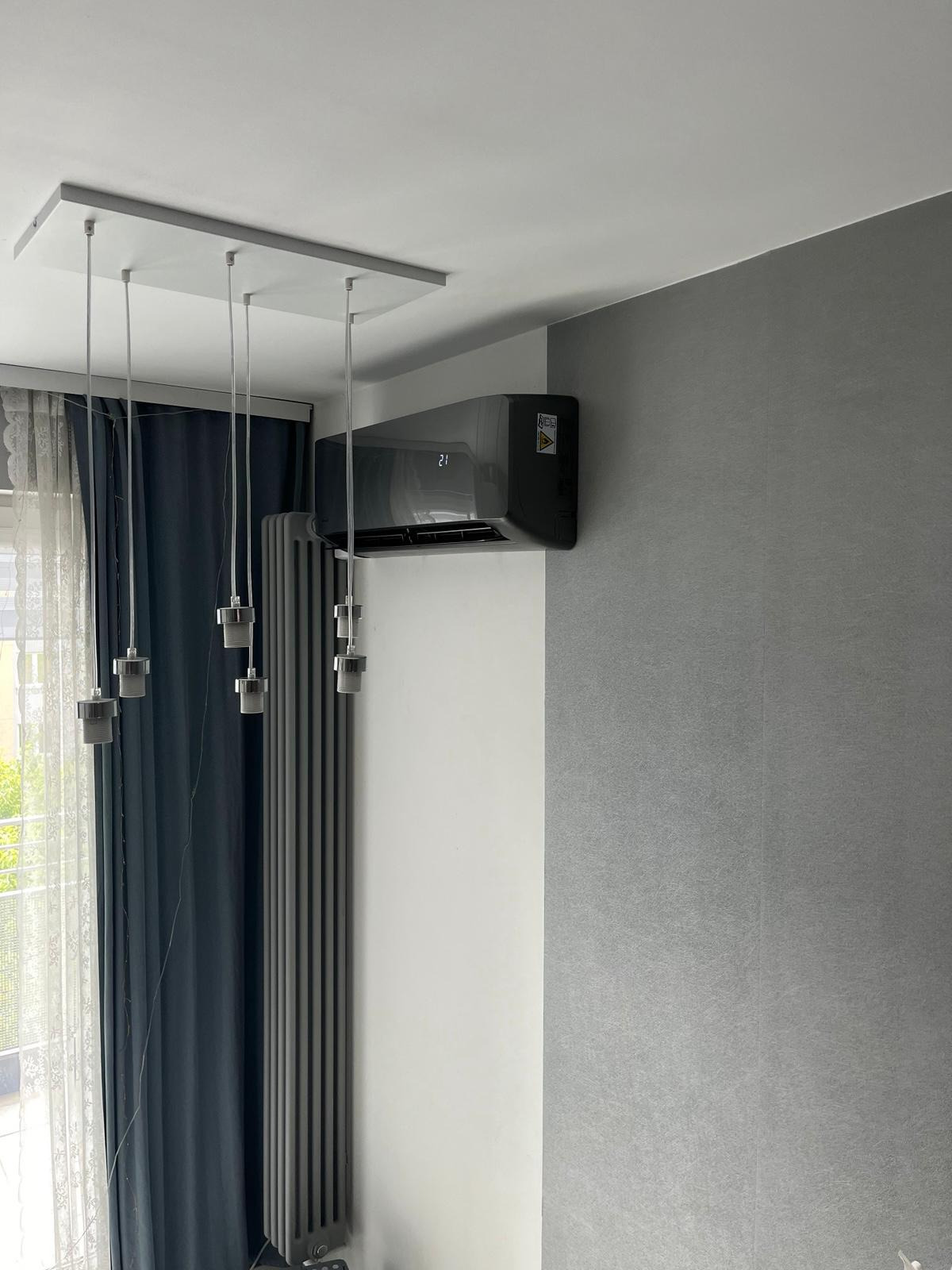 Nowoczesny, czarny klimatyzator ścienny z wyświetlaczem temperatury, zamontowany w szarym pokoju z grzejnikiem i lampą wiszącą. Widok z dołu.