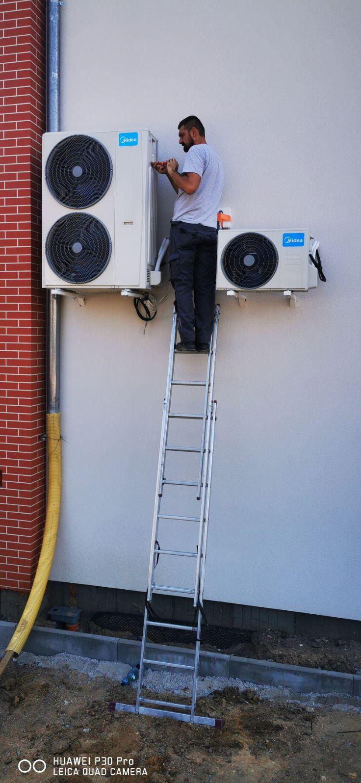Instalator na drabinie montuje dwie jednostki klimatyzacji Midea na elewacji budynku z ceglanym narożnikiem. Widoczne przewody i elementy montażowe.