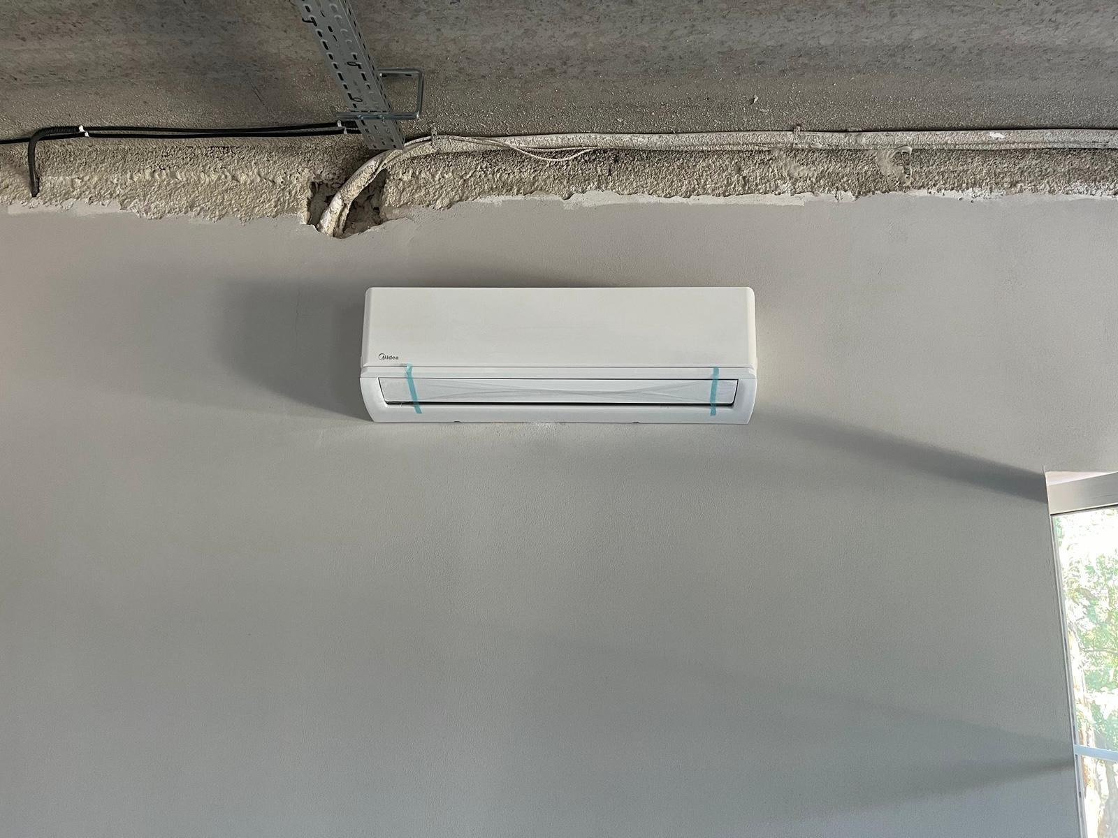 Biała klimatyzacja Midea na szarej ścianie, zabezpieczona folią ochronną. Widoczne kable i instalacja pod sufitem w surowym stanie. Minimalistyczny design.