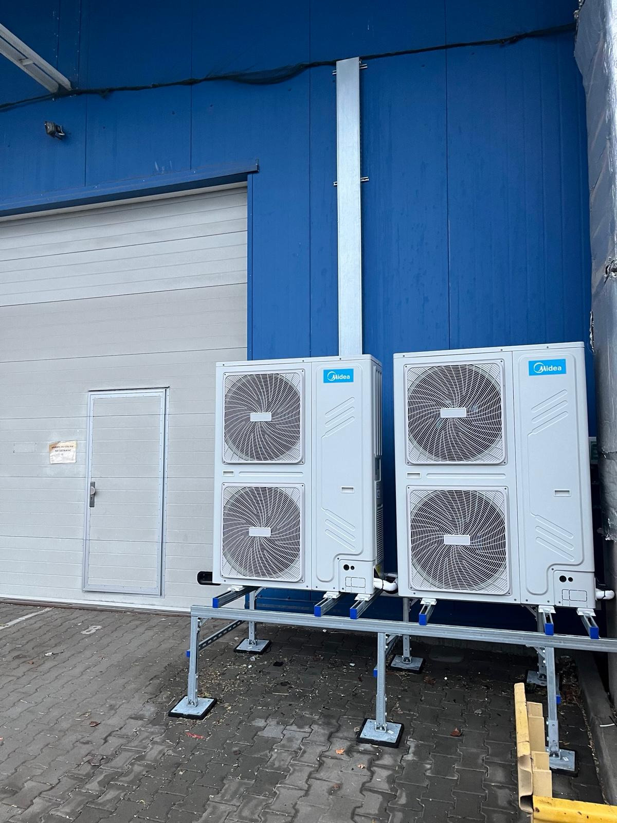 Dwa białe klimatyzatory Midea zamontowane na metalowej konstrukcji przed niebiesko-białym budynkiem. Widoczne rury i kostka brukowa. Chłodne oświetlenie.