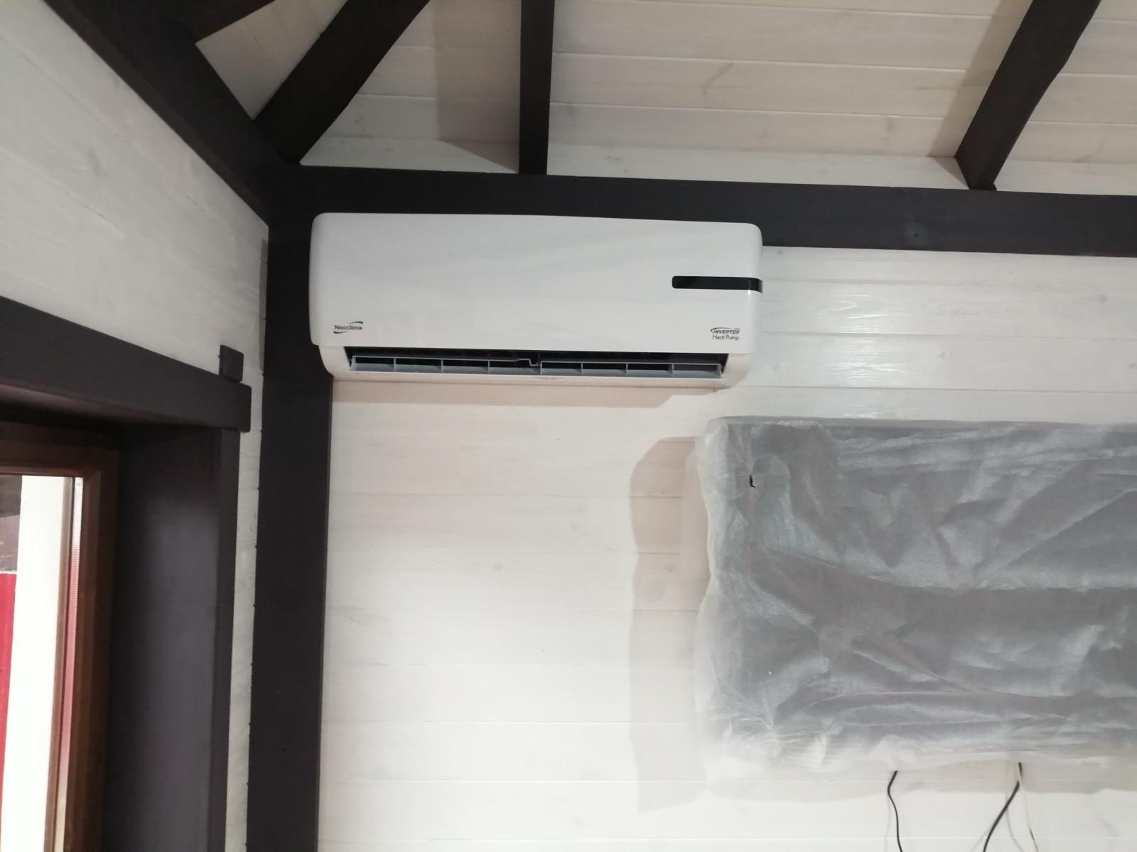 Biały klimatyzator Neoclima zamontowany na ścianie z boazerii w pomieszczeniu z drewnianą konstrukcją stropu. Obok folia ochronna na ścianie.