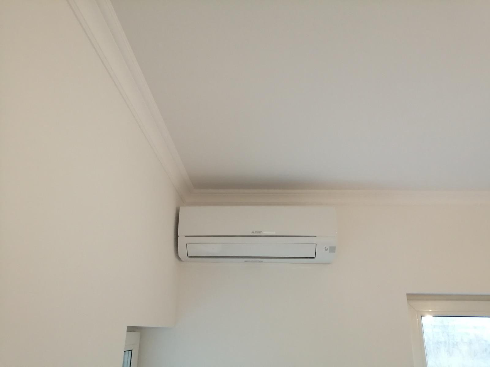 Biały klimatyzator Mitsubishi zamontowany na jasnej ścianie w rogu pokoju z listwą przysufitową i fragmentem okna. Minimalistyczny design.