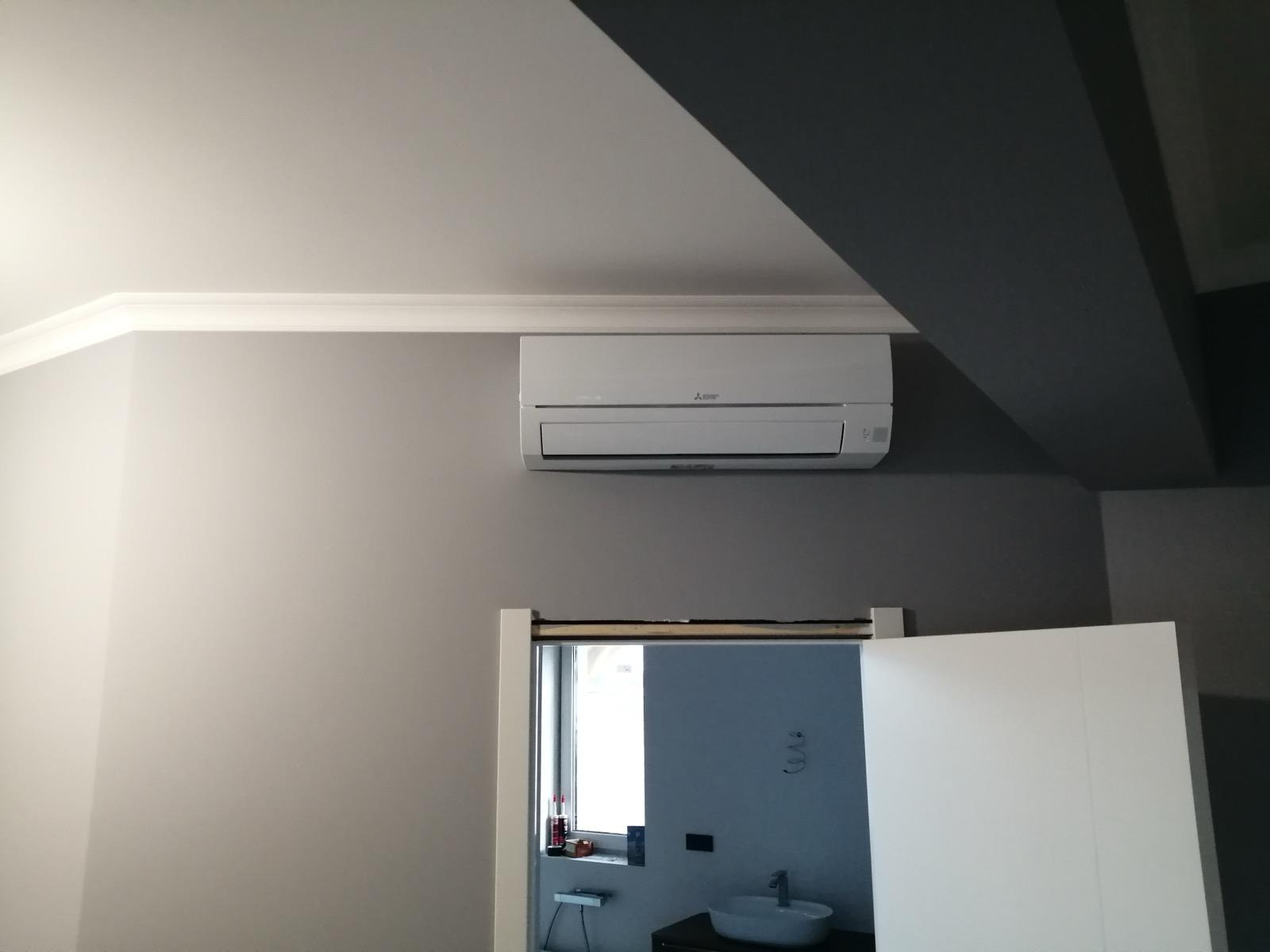 Biały klimatyzator Mitsubishi zamontowany na szarej ścianie nad drzwiami do łazienki z widoczną umywalką i oknem. Minimalistyczny design wnętrza.