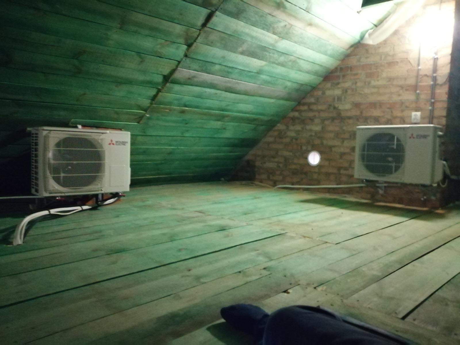 Dwa klimatyzatory Mitsubishi zamontowane na poddaszu z zieloną drewnianą podłogą i ścianami z cegły. Widoczne przewody i instalacja elektryczna.