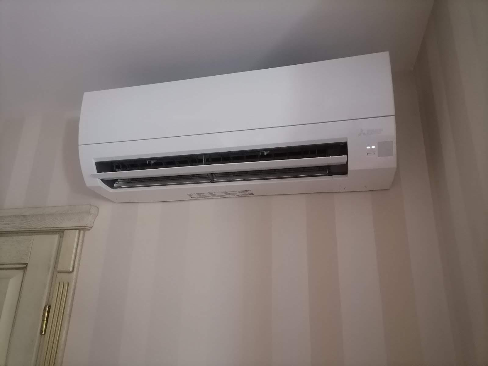 Biała klimatyzacja Mitsubishi zamontowana na ścianie z delikatnymi pionowymi paskami, widoczny fragment ozdobnej szafki po lewej stronie.
