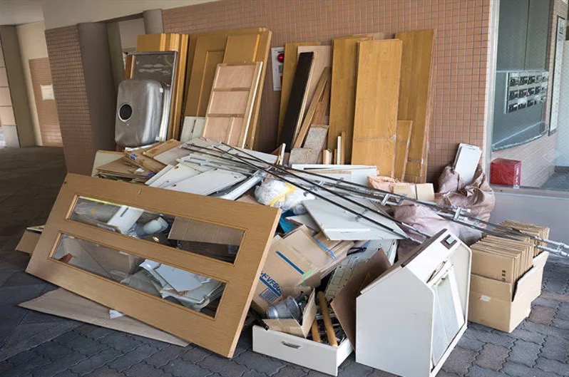 Stos różnorodnych odpadów budowlanych i meblowych, w tym drzwi, rury i kartony, zgromadzony w korytarzu budynku z brązową mozaiką na ścianie.