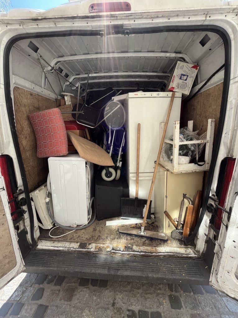 Wnętrze białego busa wypełnione różnorodnymi przedmiotami, w tym pralką, lodówką, krzesłem i kartonami, sugerujące przeprowadzkę lub opróżnianie mieszkania.