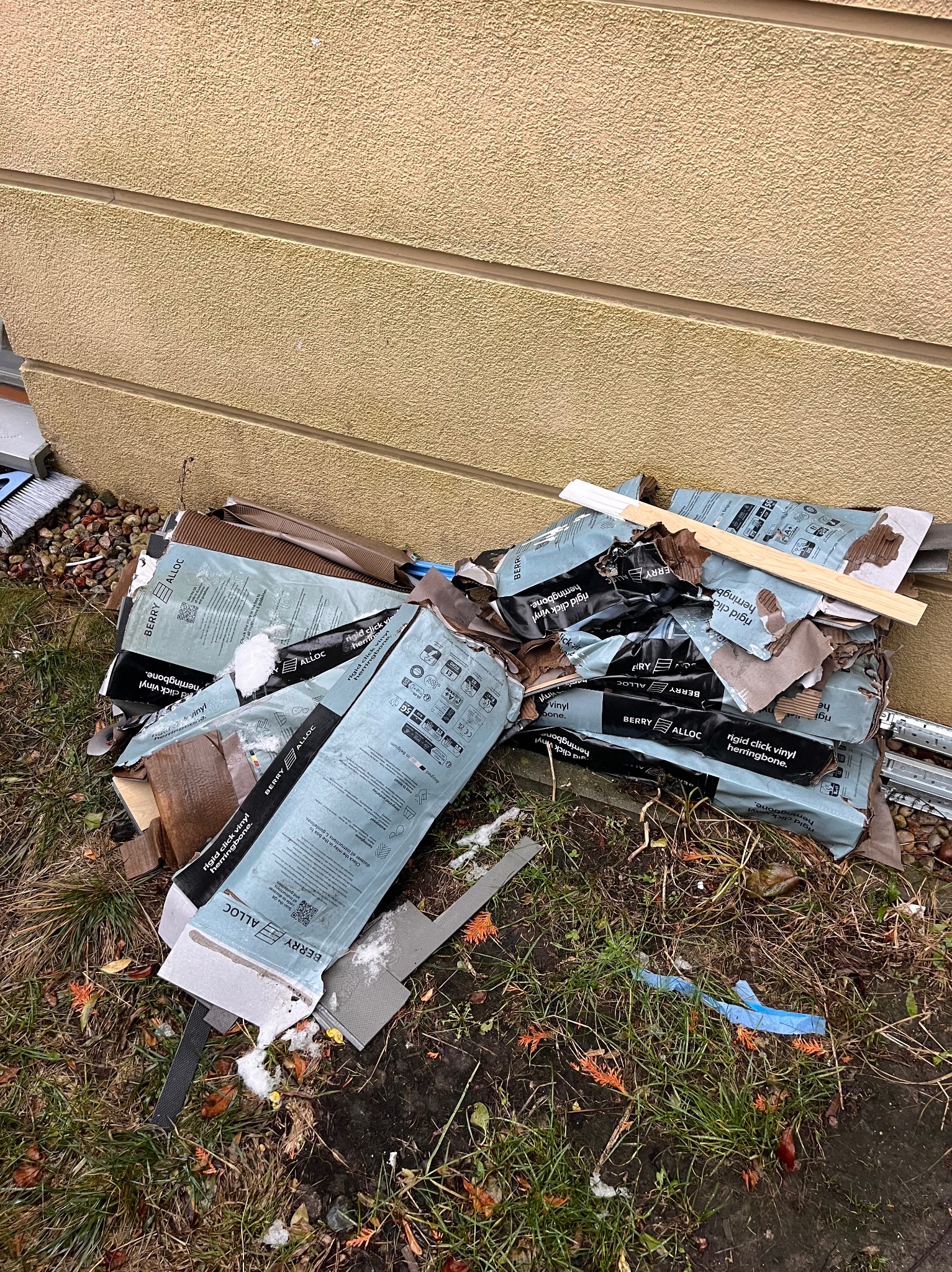 Stos zniszczonych kartonów po panelach podłogowych Berry Alloc leży na trawie przy ścianie budynku. Widoczne śmieci i resztki po remoncie.