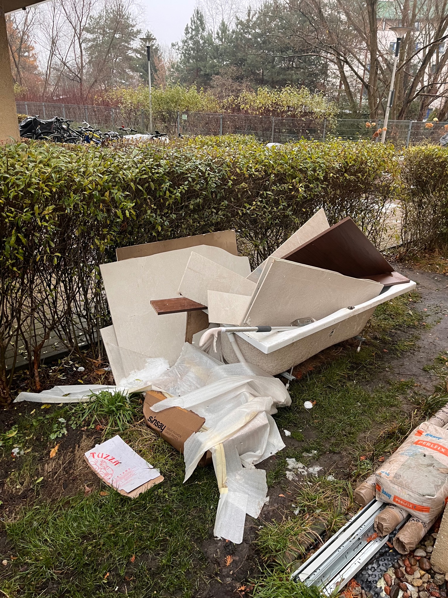 Stara wanna wypełniona gruzem i płytkami, porzucona na trawniku przed budynkiem. Widoczne worki z zaprawą i resztki opakowań. Tło stanowią krzewy i drzewa.