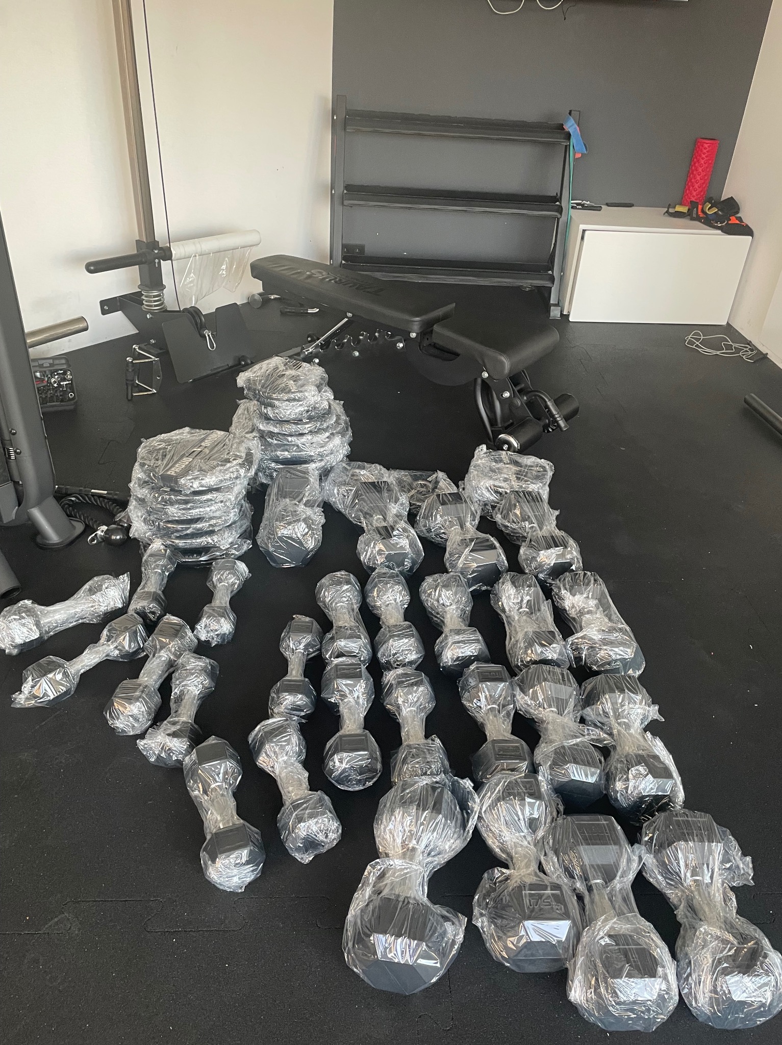 Sprzęt do ćwiczeń siłowych, hantle i obciążenia w folii, ułożone na czarnej macie w siłowni. Widoczna ławka do ćwiczeń i regał na akcesoria.