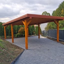 Jantar - Drewniana wiata garażowa z czerwonym dachem na posesji z kostką brukową. Widoczne słupy, konstrukcja dachu i fragment ogrodu z tujami. Ujęcie z niskiej perspektywy.