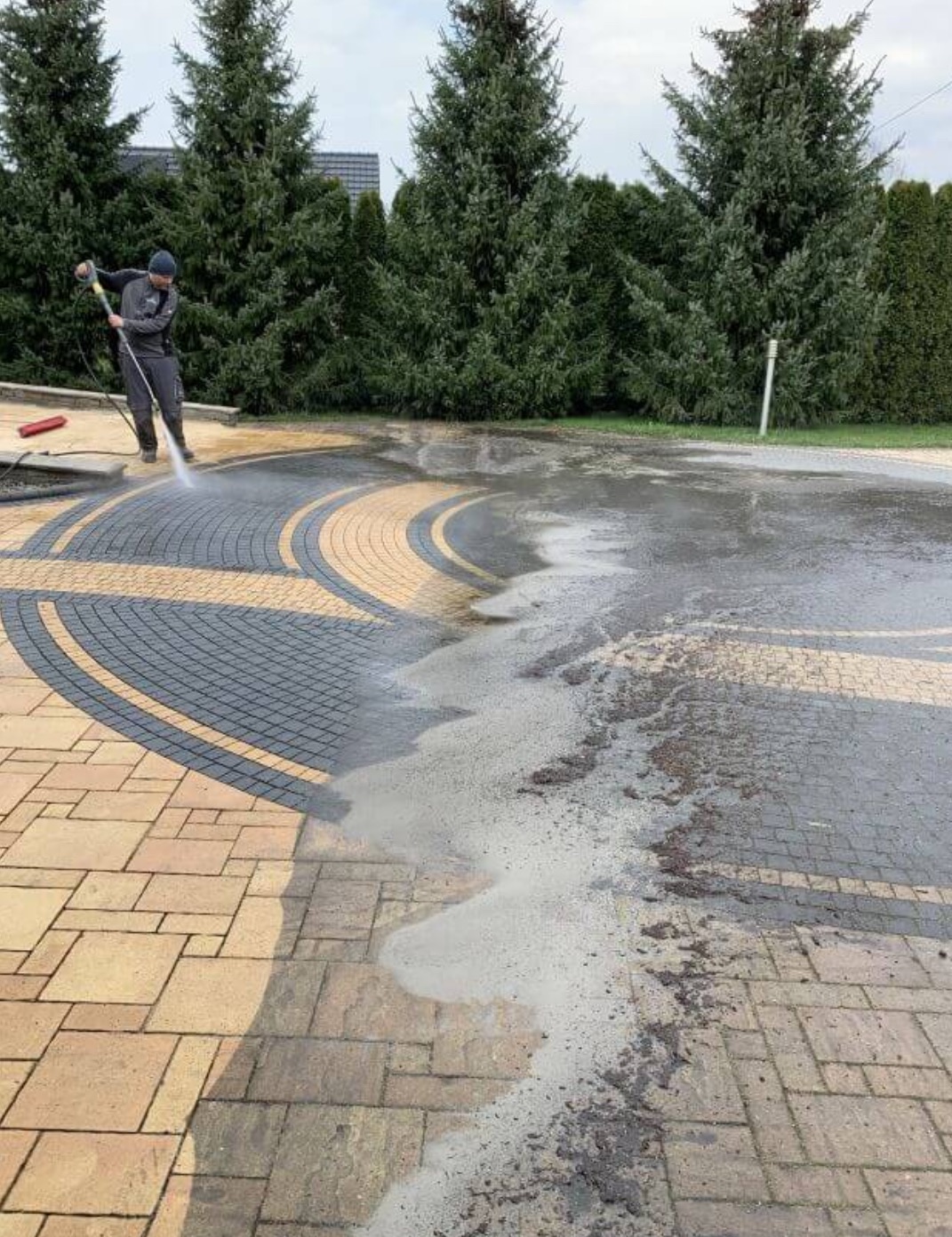 Czyszczenie kostki brukowej myjką ciśnieniową. Widoczna różnica między umytą a brudną powierzchnią. Osoba w kombinezonie obsługuje urządzenie.