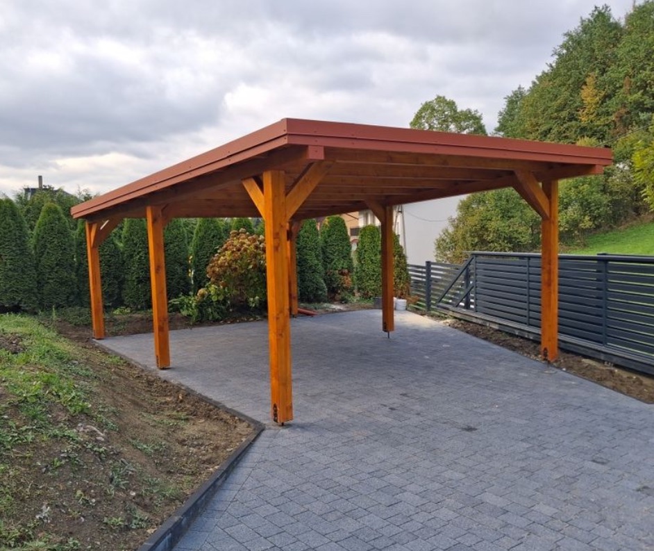 Drewniana wiata garażowa z czerwonym dachem na posesji z kostką brukową. Widoczne słupy, konstrukcja dachu i fragment ogrodu z tujami. Ujęcie z niskiej perspektywy.