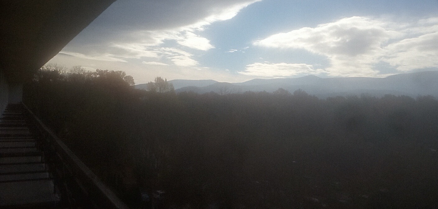 Widok z balkonu na zalesione wzgórza w oddali, przesłonięte mgłą. Schody po lewej stronie kadru. Pochmurne niebo z prześwitami błękitu.