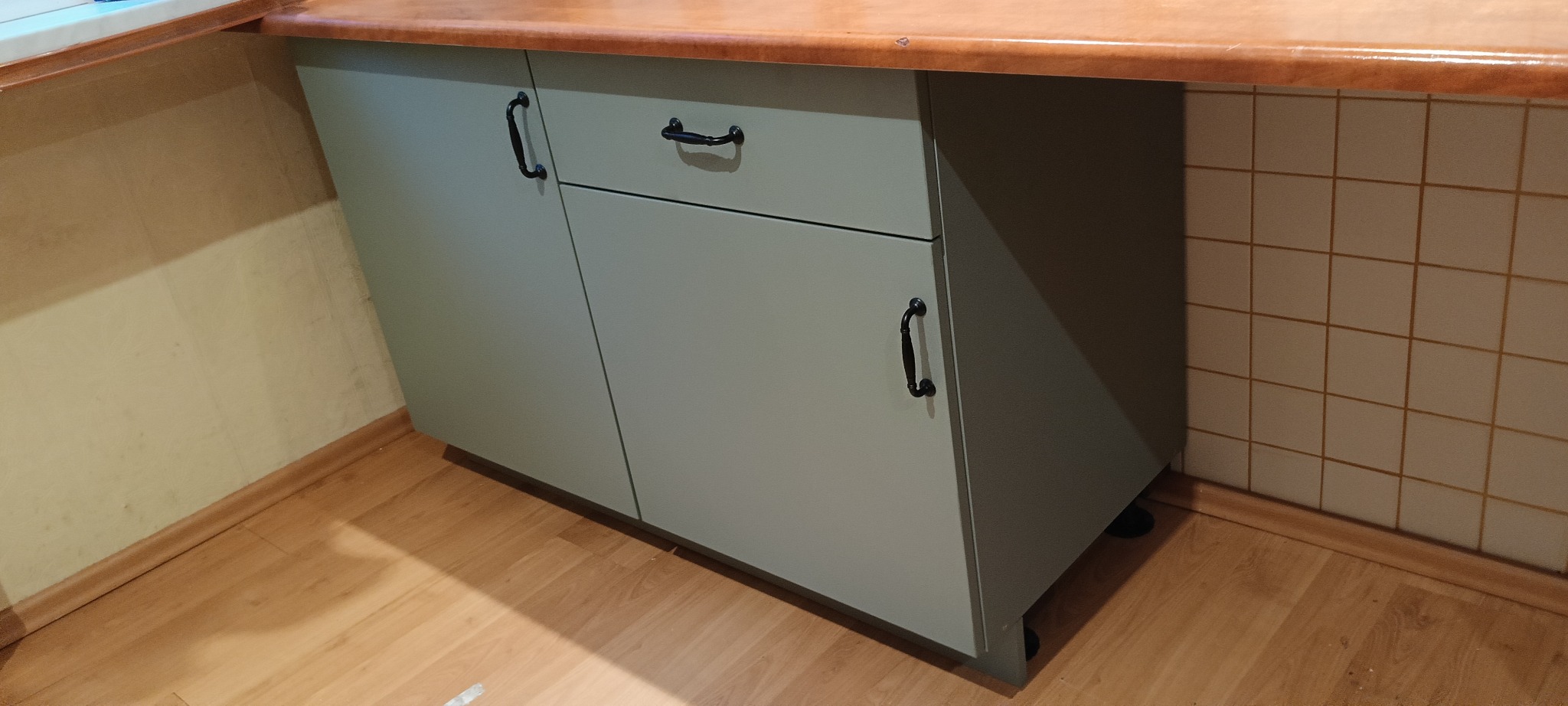 Szara szafka kuchenna z blatem w kolorze drewna, czarne uchwyty, ustawiona na podłodze z paneli, widoczna część ściany z kwadratowymi płytkami.