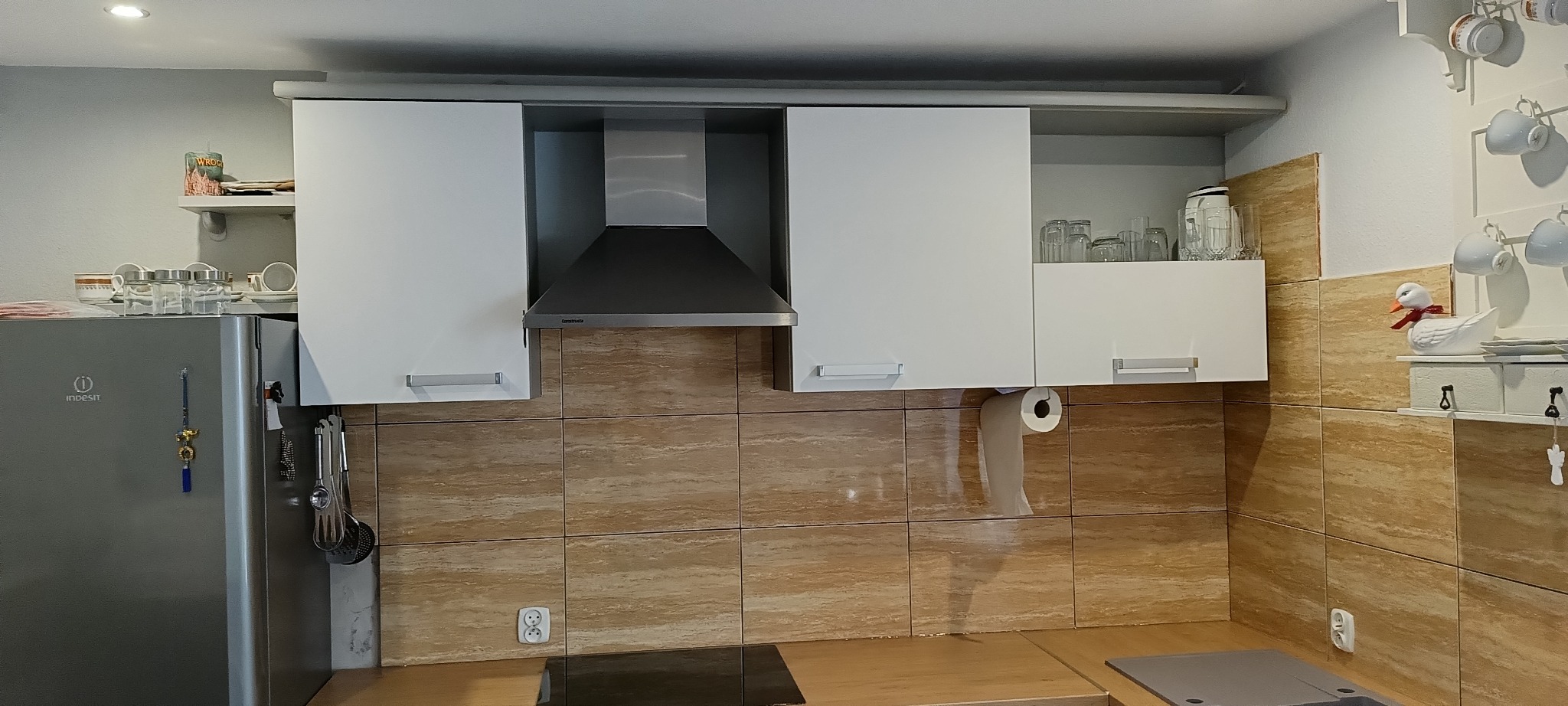 Białe szafki kuchenne z okapem i brązowymi płytkami w Pyskowicach. Widoczne naczynia i akcesoria kuchenne. Wymiana frontów i blatów.