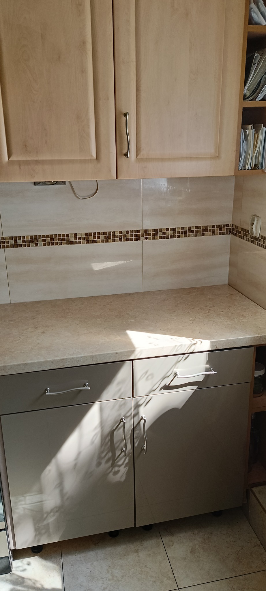 Jasne, drewniane szafki kuchenne z metalowymi uchwytami, beżowy blat i mozaikowe płytki. Na szafkach cień od promieni słonecznych.